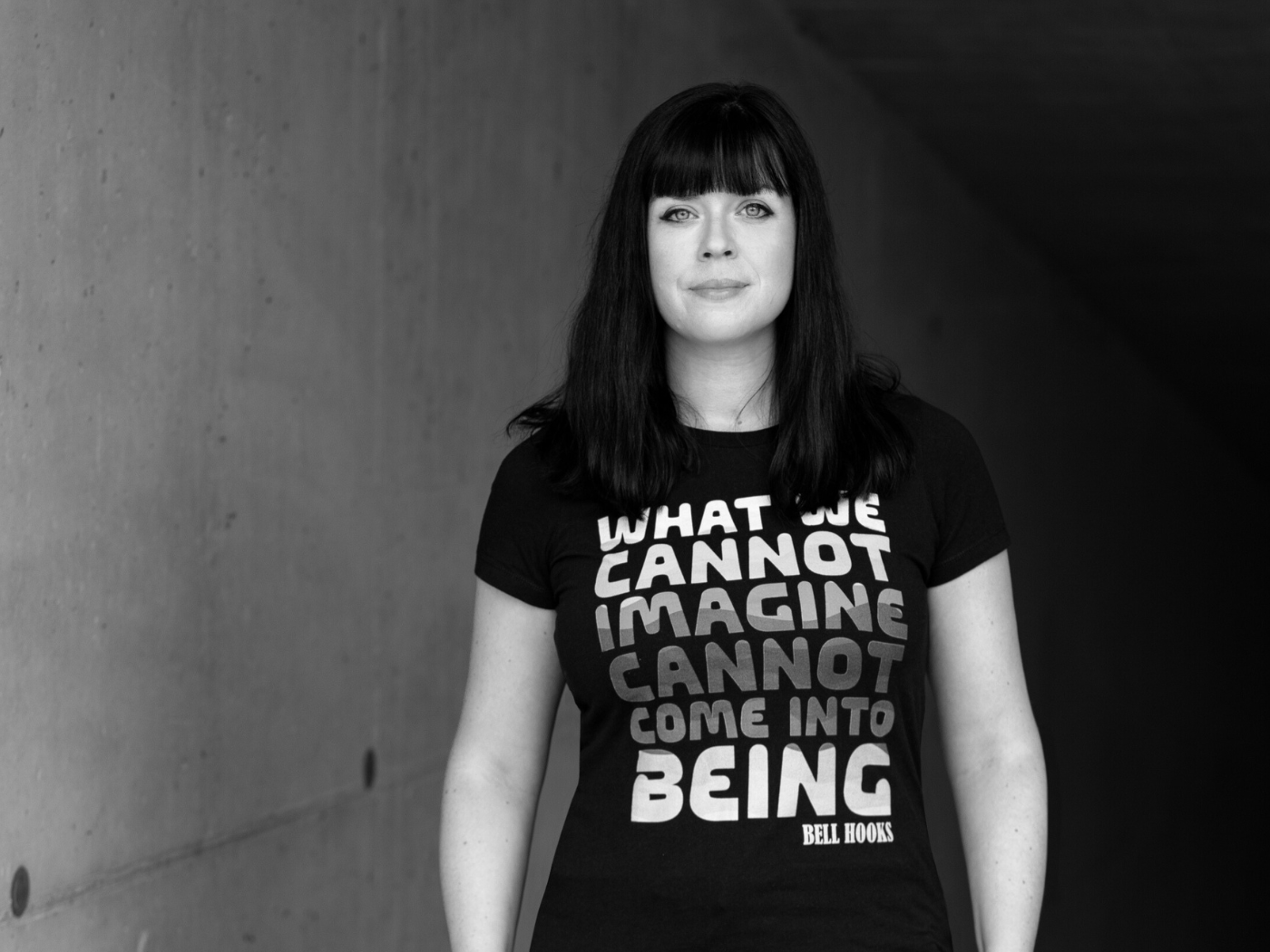Ein Portrait der Autorin Mareike Fallwickl. Sie schaut ernst, aber freundlich in die Kamera und trägt ein Tshirt mit dem Aufdruck eines bell hooks Zitats "What we cannot imagine cannot come into being"